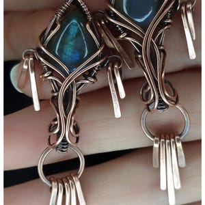 May include: A pair of handcrafted earrings featuring a central, iridescent blue-green gemstone encased in intricate copper wire. The earrings have a geometric design with dangling metal accents and the text "KELLY J JEWELLERY".