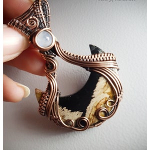 K&ouml;nnte beinhalten: Ein Anh&auml;nger aus Kupferdraht, der einen sichelf&ouml;rmigen schwarzen und wei&szlig;en Achat-Stein mit einem kleineren wei&szlig;en Mondstein an der Spitze umwickelt. Der Anh&auml;nger ist handgefertigt und hat ein filigranes, kompliziertes Design.