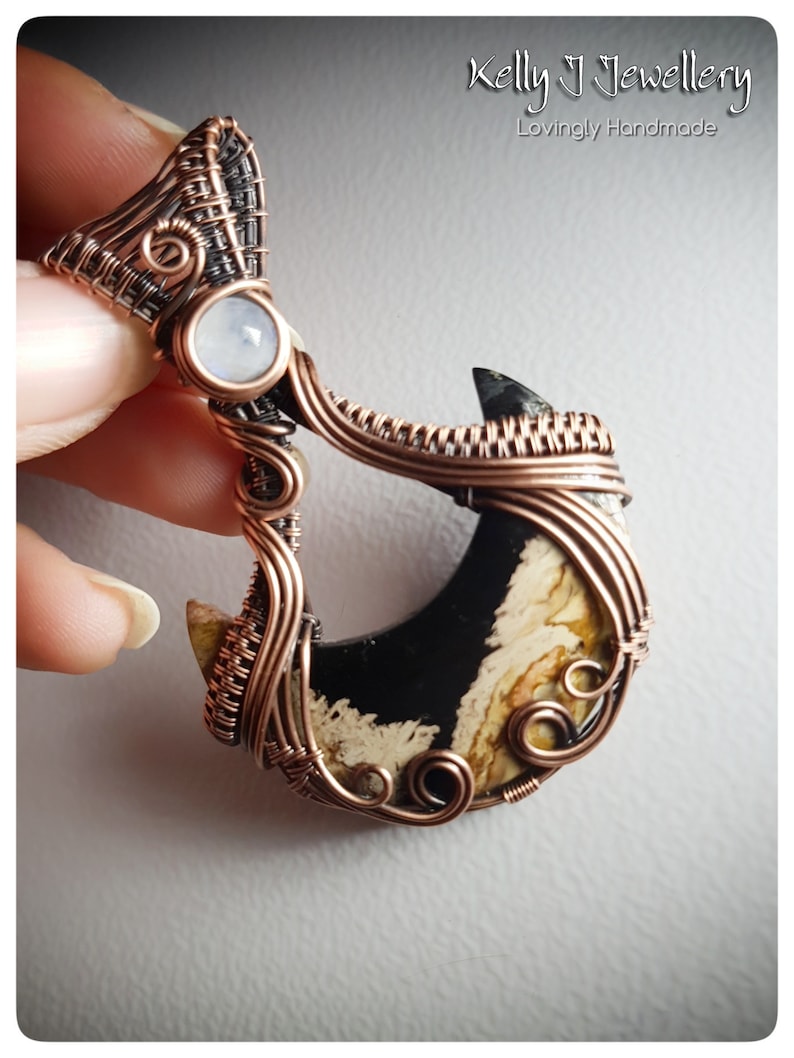 K&ouml;nnte beinhalten: Ein Anh&auml;nger aus Kupferdraht, der einen sichelf&ouml;rmigen schwarzen und wei&szlig;en Achat-Stein mit einem kleineren wei&szlig;en Mondstein an der Spitze umwickelt. Der Anh&auml;nger ist handgefertigt und hat ein filigranes, kompliziertes Design.