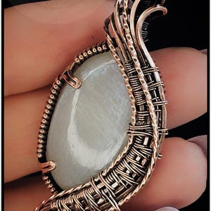 May include: A pendant featuring an oval, translucent white moonstone cabochon encased in intricate, oxidized copper wire wrapping. The metalwork consists of twisted and straight wire coils forming a decorative frame with a top bail loop.