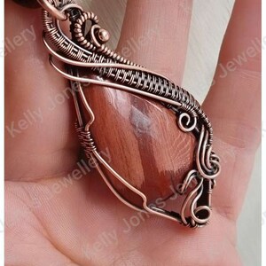 May include: A copper wire wrapped pendant featuring a red and brown gemstone. The pendant is hanging from a brown cord.