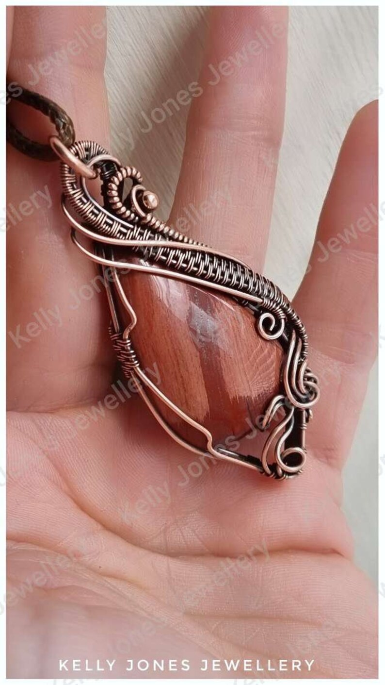 May include: A copper wire wrapped pendant featuring a red and brown gemstone. The pendant is hanging from a brown cord.