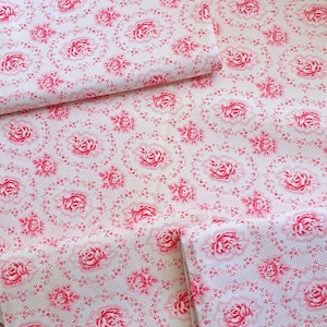 Könnte beinhalten: Weißer Stoff mit einem sich wiederholenden Muster aus rosa Rosen und kleinen floralen Akzenten. Die Rosen sind von einem gepunkteten Rand umgeben, was eine Vintage- und romantische Ästhetik erzeugt. Der Stoff ist an mehreren Stellen gefaltet.