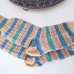 Könnte beinhalten: Ein Paar gestrickte Socken mit Streifen in Blau, Grün, Rosa und Beige. Die Socken sind aus weichem Garn gefertigt. Diese gemütlichen Socken sind perfekt, um die Füße warm zu halten.