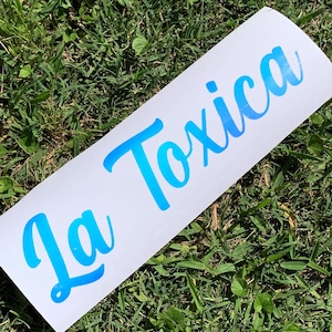 May include: A white sticker with the text "La Toxica" written in blue cursive lettering. The sticker has a gradient effect, with the lettering fading from a darker blue to a lighter blue.