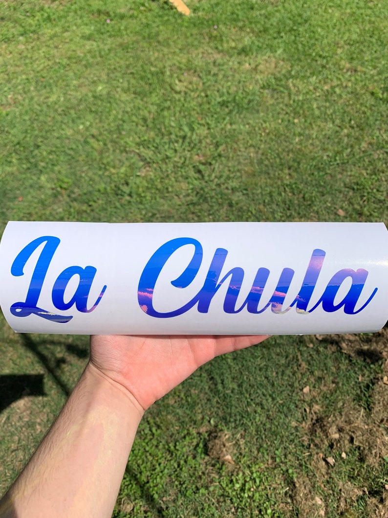 Pu&ograve; includere: Un adesivo in vinile bianco con il testo "La Chula" in lettere corsive blu. Le lettere hanno un effetto sfumato con uno schema di colori rosa e blu.