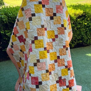 Peut inclure: Un quilt en patchwork composé d'une grille de carrés de tissu dans des tons jaune, orange, pêche et crème. La courtepointe est agrémentée de petits carrés de connexion marron foncé et présente une texture matelassée en tourbillons.
