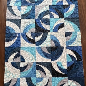 May include: A quilted blanket with a geometric design in shades of blue, grey, and white. The quilt features a repeating pattern of curved shapes, creating a modern and artistic look. The quilting adds texture and detail to the fabric.