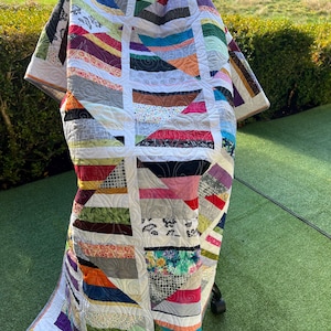 May include: A vibrant patchwork quilt featuring a mix of patterned fabrics in various colours. The quilt has a geometric design with white borders and detailed stitching. It is draped over an object on a green surface, likely a lawn.