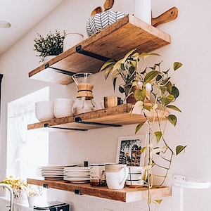 May include: Three rustic wooden shelves with black metal brackets are mounted on a white wall. The shelves display various kitchen items, including white plates, mugs, a glass coffee maker, and potted plants. The shelves are made of reclaimed wood.