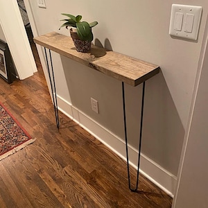 May include: A wooden console table with a natural finish, featuring a rectangular top and black hairpin legs. A potted plant sits on the table's surface. The table is approximately 48 inches long.
