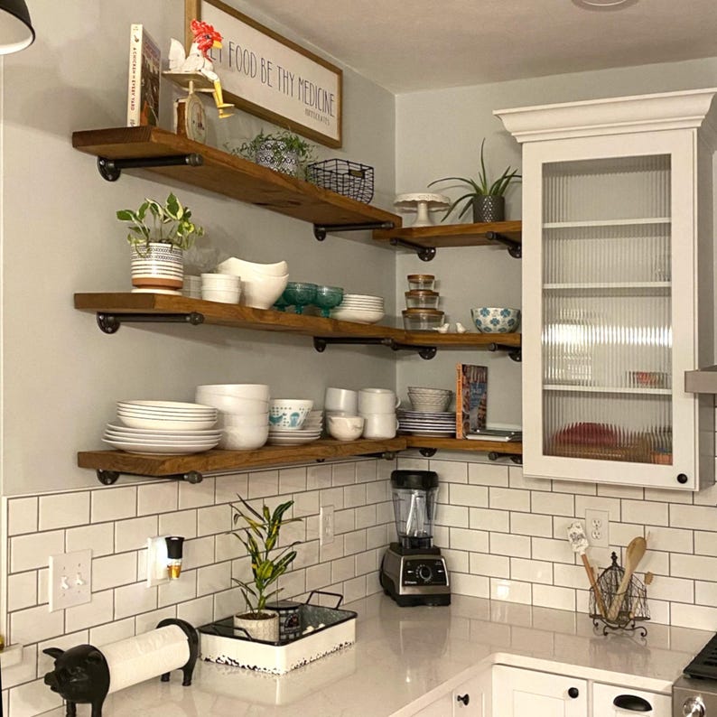 May include: Three tiered wooden floating shelves with black metal pipe brackets mounted in a kitchen corner. The shelves hold white ceramic bowls, plates, glassware, small potted plants, and a sign that reads Food Be Thy Medicine.