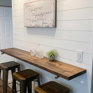 May include: A long, dark-stained wooden floating bar shelf mounted on a white shiplap wall. Below the shelf are three dark metal stools. On the shelf sit two clear glass tumblers, a glass decanter, and a small green air plant in a gray pot.