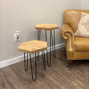 May include: Two hexagonal side tables with natural light-toned wood grain tops and thin black metal hairpin legs. The tables are of different heights, with the smaller table positioned in front of the taller one, both featuring a smooth finish.