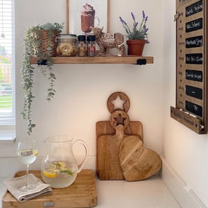 May include: A wooden shelf holds a potted plant, a glass jar, spice bottles, a cow-shaped calendar, and a lavender pot. Below, a wooden tray features a glass pitcher of lemon water, a wine glass, and three wooden cutting boards with star and heart shapes.