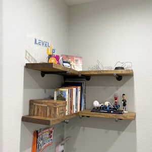 May include: Two L-shaped corner shelves made of medium-toned wood planks supported by black industrial-style metal pipe brackets. The shelves hold various items including books, a wooden box, a model race car, and a small figurine.