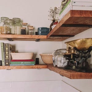 May include: Two tiers of rustic wooden corner shelves mounted with black metal brackets. The shelves hold glass storage jars, cookbooks including Salt Fat Acid Heat and Jamie Oliver Ingredients, a beige mixing bowl, and a vintage brass kitchen scale.