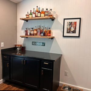 May include: A home bar setup featuring a black cabinet with gold hardware, two wooden shelves with black metal pipe brackets holding various glass liquor bottles, a green Bourbon St sign, and a framed print of a glass of whiskey on a white wall.