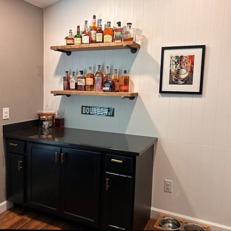 May include: A home bar setup featuring a black cabinet with gold hardware, two wooden shelves with black metal pipe brackets holding various glass liquor bottles, a green Bourbon St sign, and a framed print of a glass of whiskey on a white wall.