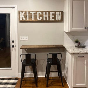 Puede incluir: Una escena de cocina con un letrero de madera con la palabra "KITCHEN" en letras blancas. Debajo del letrero, una estanter&iacute;a de madera y dos taburetes de bar de metal negro. Armarios blancos y una encimera de granito completan la escena.