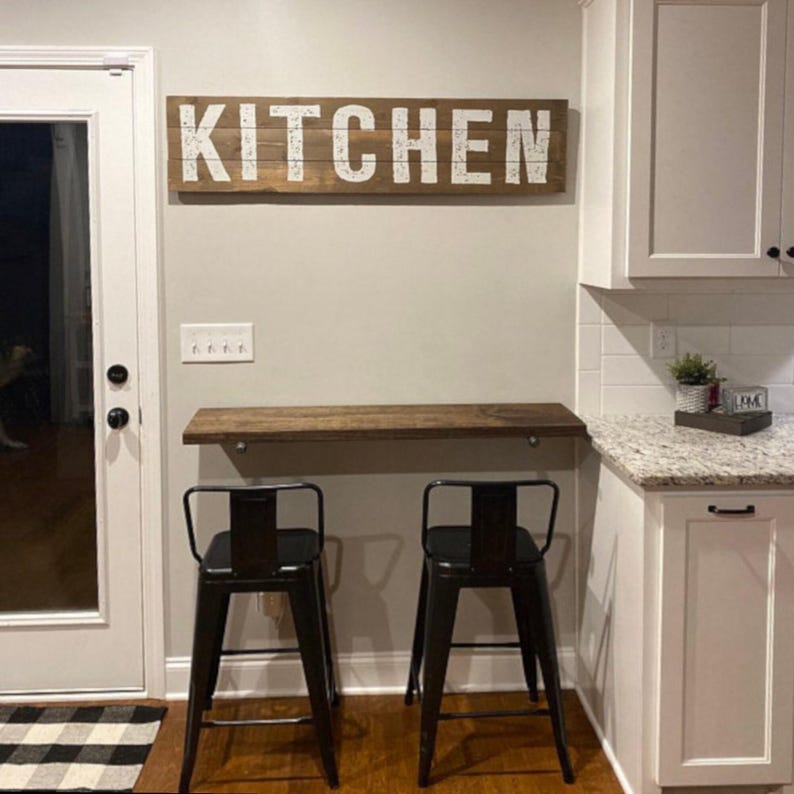 Puede incluir: Una escena de cocina con un letrero de madera con la palabra "KITCHEN" en letras blancas. Debajo del letrero, una estanter&iacute;a de madera y dos taburetes de bar de metal negro. Armarios blancos y una encimera de granito completan la escena.