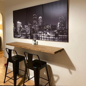 May include: A triptych canvas wall art set featuring a black and white nighttime cityscape of skyscrapers and city lights reflected in water, mounted above a long, narrow, natural-edge wooden bar table with two black metal bar stools.