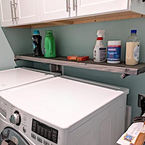 May include: A dark gray wooden shelf mounted above a white washing machine, holding a black detergent bottle, a green detergent bottle, a small orange box, a white spray bottle, a container of DisinfeX wipes, and a clear bottle with beige liquid.
