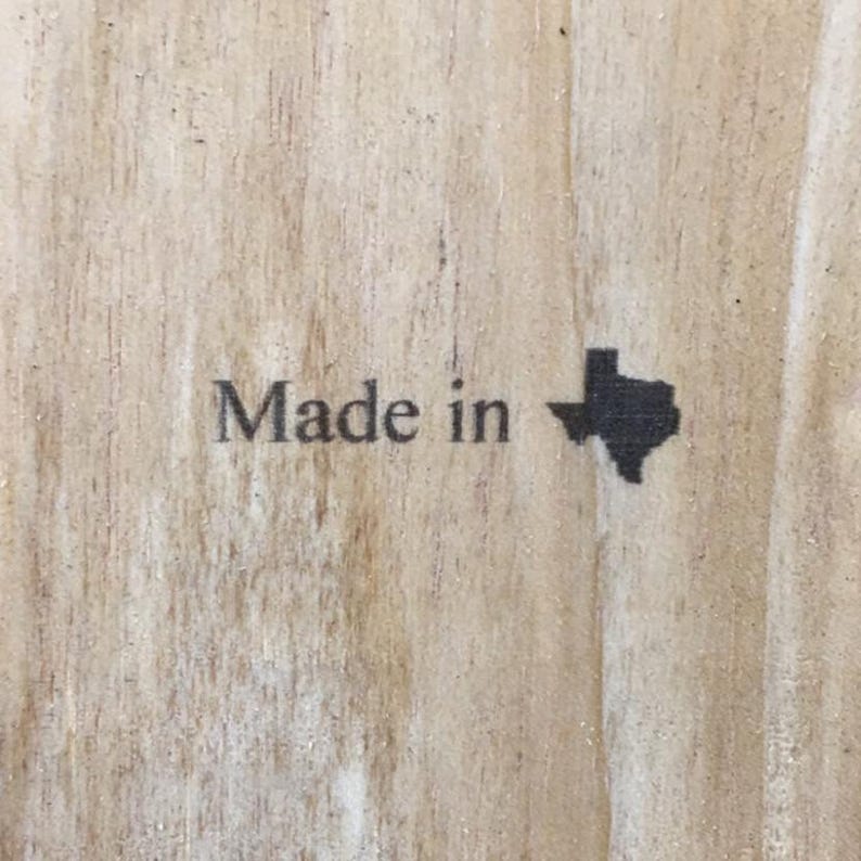 May include: A light-colored wooden surface featuring the black printed text Made in followed by a solid black silhouette of the state of Texas. The wood grain is visible throughout the background, showing natural vertical lines and light brown tones.
