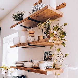 May include: Three rustic wooden shelves mounted on a white wall, displaying kitchen items. The shelves hold white plates, mugs, bowls, a glass coffee maker, and potted plants. Black metal brackets support the shelves, creating a farmhouse aesthetic.