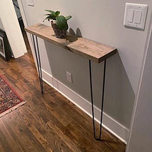 May include: A wooden console table with a natural finish, supported by black hairpin legs. A small potted plant sits on the tabletop. The table is approximately 48 inches long.