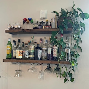 May include: Two dark wood floating shelves mounted on a wall, holding various glass liquor bottles, a metal cocktail shaker, and several stemmed glasses hanging from the bottom shelf. A trailing green pothos plant sits on the top right shelf.