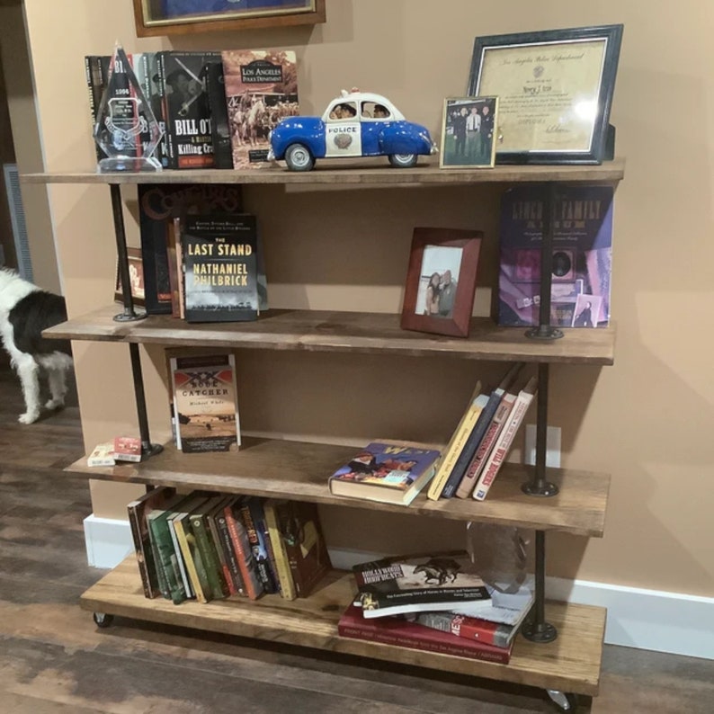 May include: A rustic, four-tiered bookshelf made of reclaimed wood and black metal pipes. The shelves are filled with books, framed photos, and decorative items, including a blue and white police car toy. The bookshelf is on wheels.