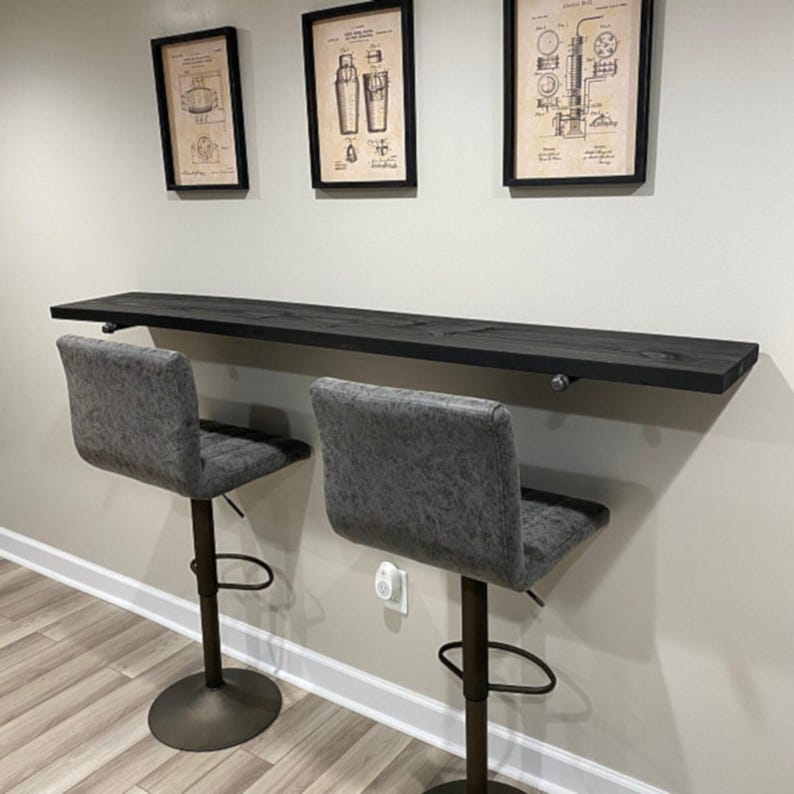 Puede incluir: Una encimera de bar de madera te&ntilde;ida oscura montada en una pared, con dos taburetes de bar tapizados en gris. Tres diagramas vintage enmarcados cuelgan sobre la barra. Los taburetes tienen altura ajustable y una base circular. El bar es de dise&ntilde;o sencillo y moderno.