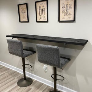 May include: A dark stained wooden bar top mounted on a wall, with two gray upholstered bar stools. Above the bar are three framed vintage patent prints. The stools have adjustable height and a circular base.