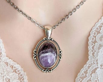 May include: A silver-toned necklace featuring an oval pendant with a polished purple and white banded amethyst stone. The stone is set within an ornate, filigree-style metal frame with decorative scrollwork details, suspended from a simple silver chain.