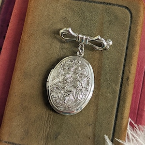 Floral embossed oval shiny silver locket with bow style pin displayed on a stack of vintage books.