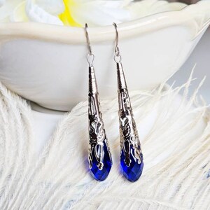 Vintage Style earrings with long silver tone filigree bead caps over sapphire blue faceted glass beads. Shown dangling from a bowl.