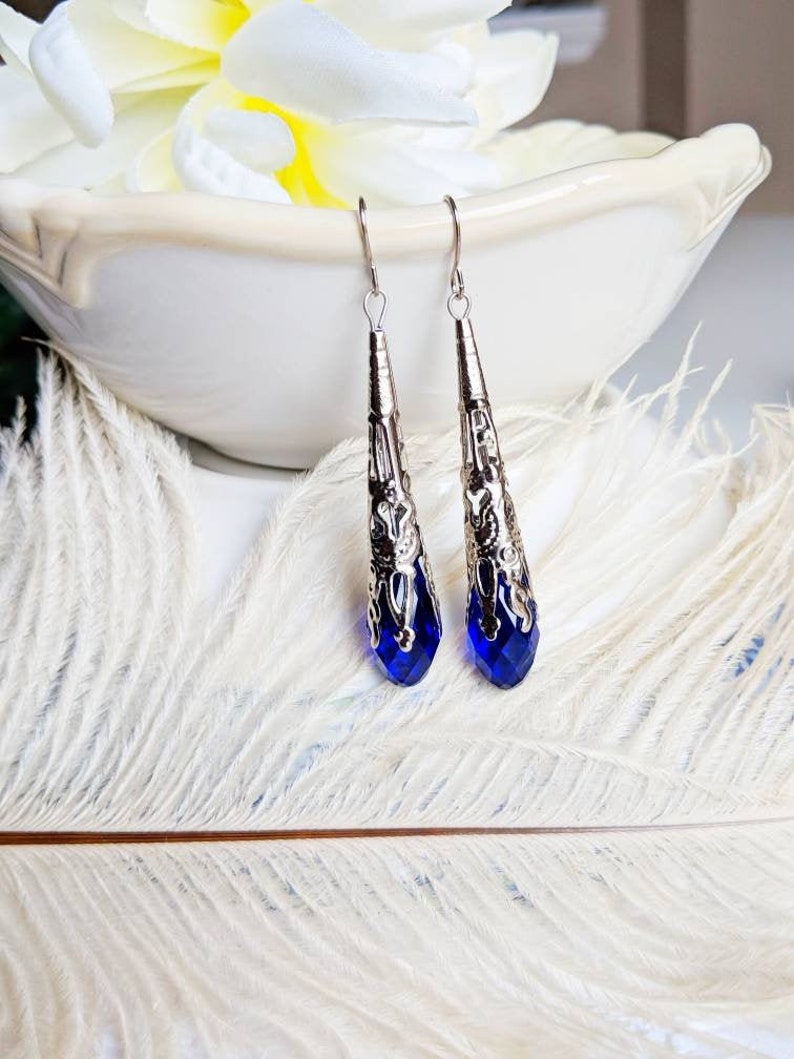 Vintage Style earrings with long silver tone filigree bead caps over sapphire blue faceted glass beads. Shown dangling from a bowl.