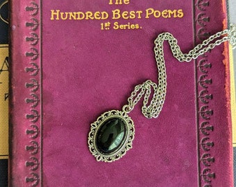 May include: A silver-toned necklace featuring an oval pendant with a smooth black stone set in an intricate filigree frame. The pendant rests on a thin silver chain, displayed against the cover of a purple book titled The Hundred Best Poems 1st Series.