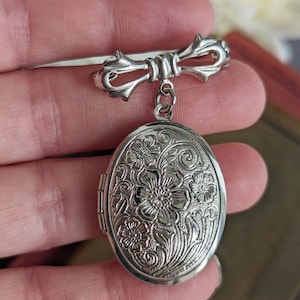 Floral embossed oval shiny silver locket with bow style pin shown being held in a hand.