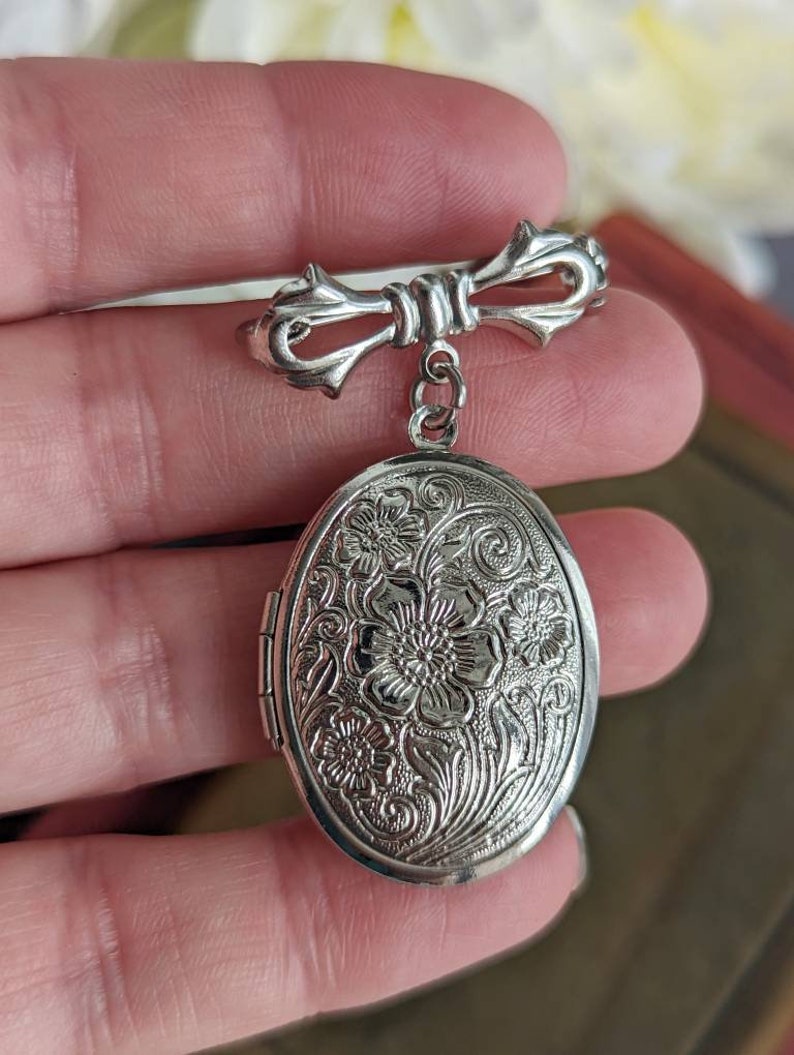 Floral embossed oval shiny silver locket with bow style pin shown being held in a hand.