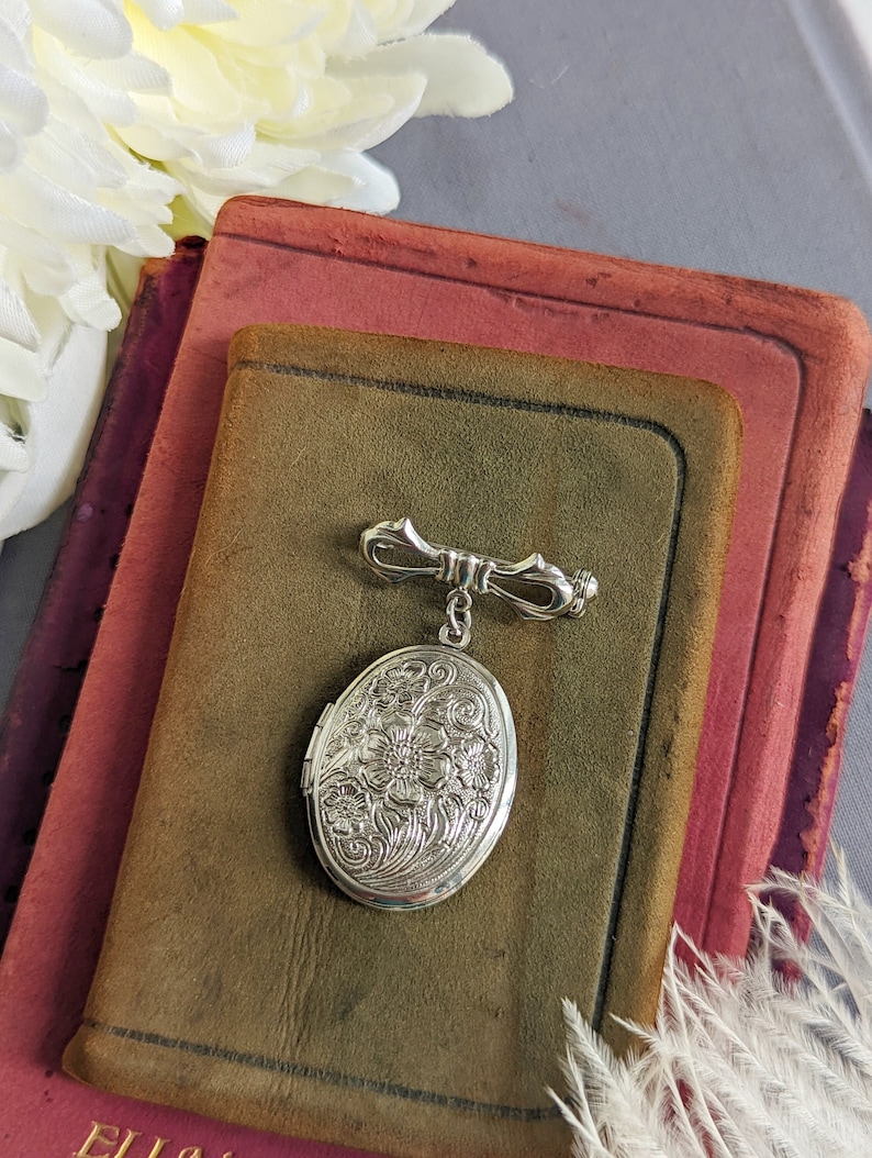 Floral embossed oval shiny silver locket with bow style pin displayed on a stack of vintage books.