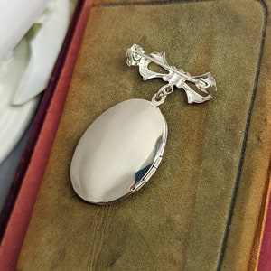 Back of floral embossed oval shiny silver locket with bow style pin displayed on a stack of vintage books.