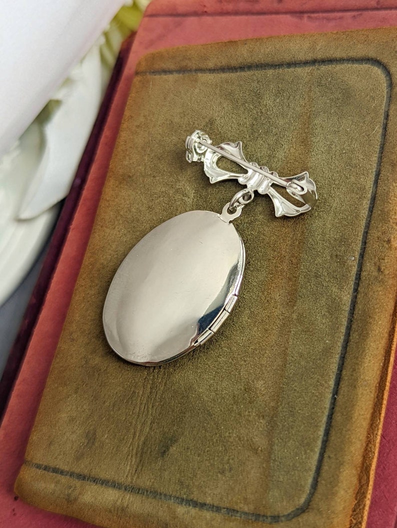 Back of floral embossed oval shiny silver locket with bow style pin displayed on a stack of vintage books.