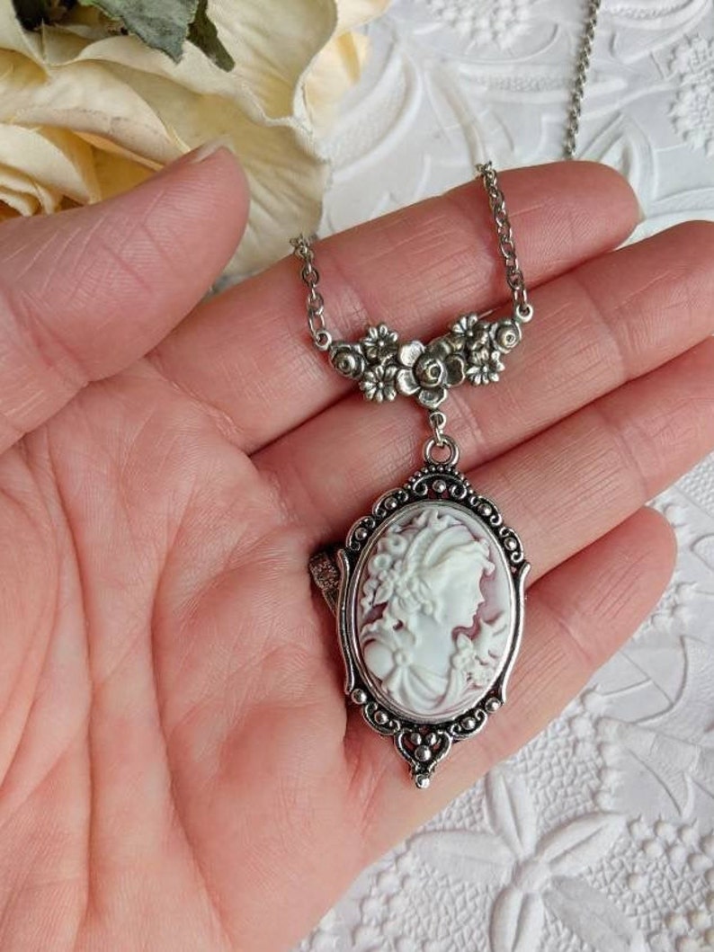 Hand holding cameo necklace in antiqued silver finish. Cameo features a Grecian Goddess with a small bird. The cameo colour is white on a wine background.