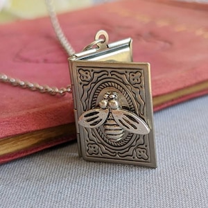 May include: A silver locket necklace featuring a bee design. The locket is open and has an intricate design around the bee.