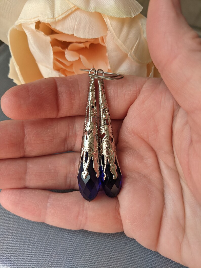 Vintage Style earrings with long silver tone filigree bead caps over sapphire blue faceted glass beads. Shown being held in a hand.