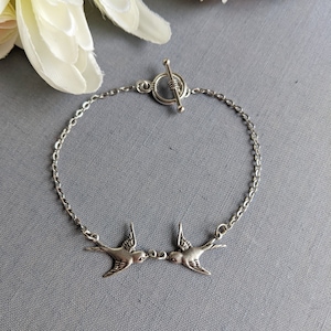 Peut inclure: Bracelet argenté orné de deux oiseaux se faisant face. La chaîne est fine et le fermoir est de type toggle. Les oiseaux sont détaillés avec des ailes et des becs. Le fond est gris uni. Longueur approximative 18 cm.