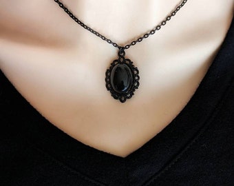 May include: A black necklace featuring a central oval-shaped black cabochon stone set within an intricate, scalloped metal frame. The pendant hangs from a delicate black chain, creating a monochromatic piece of jewelry with a detailed border.