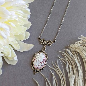 Ornate silver Cameo necklace with white and wine cameo displayed on grey background with flowers and feathers surrounding it.